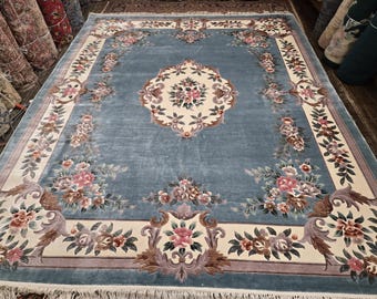 May include: A large, rectangular area rug with a light blue field and an ornate floral design. The rug features a central medallion and a border with cream, pink, and brown floral motifs. The rug is made of a soft material and has a fringe.