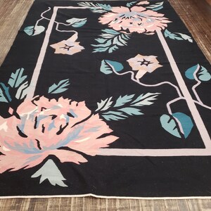May include: A black rug with a pink and blue floral pattern. The rug has a white border and is framed by a light pink rectangle.