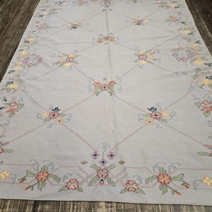 May include: A rectangular, light blue rug with a floral pattern. The rug features a repeating design of flowers in shades of pink, yellow, and green, outlined with a light pink border. The rug is placed on a dark wood floor.