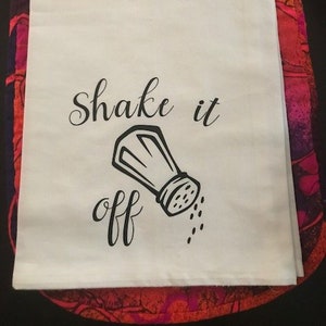 May include: White kitchen towel with black text that reads "Shake it off" and a black and white illustration of a salt shaker pouring salt.