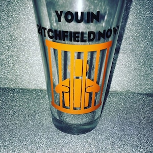 May include: A clear glass with an orange graphic of a person in a jail cell. The text "YOU IN BITCHFIELD NOW!" is printed on the glass.