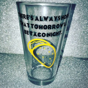 May include: A clear glass with a black and yellow design that says "There's always hope that tomorrow'll be taco night."  A yellow taco is pictured on the glass.