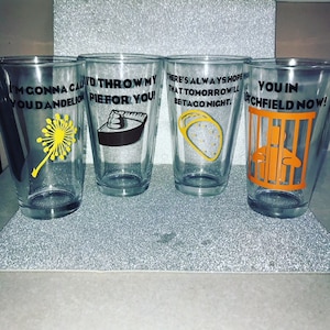 May include: Four clear glass cups with black text and colorful graphics. The text on the cups reads "I'm gonna call you dandelion", "I'd throw my pie for you", "There's always hope that tomorrow'll be taco night", and "You in Litchfield now!"