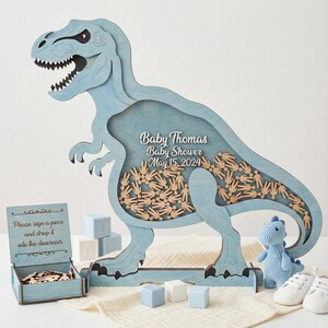 May include: A light blue wooden Tyrannosaurus Rex-shaped baby shower guest book. The dinosaur has a hollow center filled with wooden hand-shaped pieces. The text "Baby Thomas Baby Shower May 15, 2024" is displayed on the dinosaur. Additional items include a box with wooden pieces, blocks, and a small stuffed dinosaur.