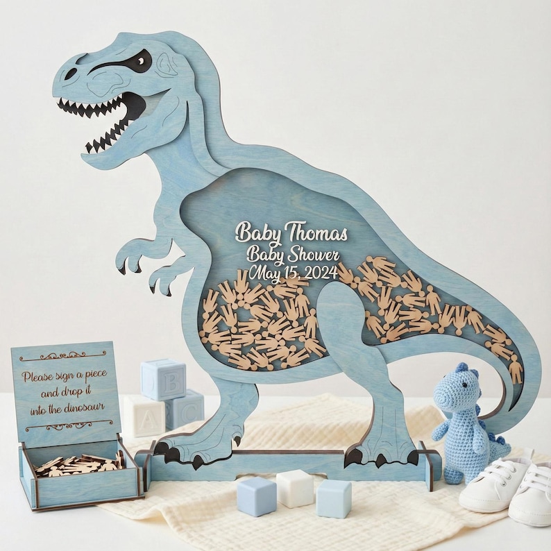 May include: A light blue wooden Tyrannosaurus Rex-shaped baby shower guest book. The dinosaur has a hollow center filled with wooden hand-shaped pieces. The text "Baby Thomas Baby Shower May 15, 2024" is displayed on the dinosaur. Additional items include a box with wooden pieces, blocks, and a small stuffed dinosaur.