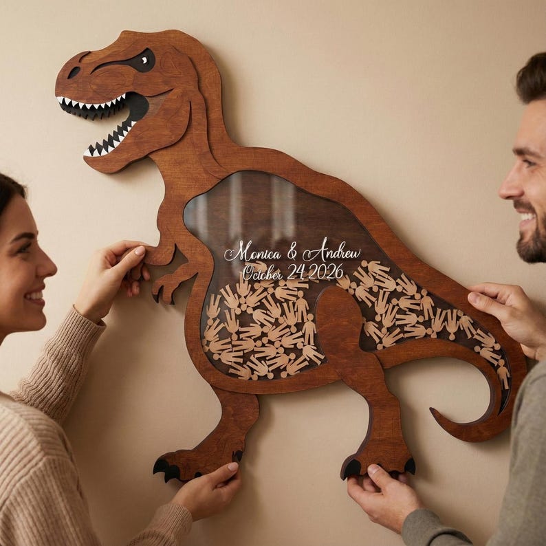 May include: A wooden Tyrannosaurus Rex-shaped guest book alternative. The brown dinosaur has a clear window filled with small wooden figures. The text "Monica & Andrew October 24 2026" is displayed on the window.