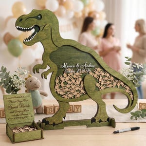 May include: A green wooden Tyrannosaurus Rex guest book alternative with a hollowed-out center filled with small wooden figures. The text "Monica & Andrew October 24 2026" is inscribed on the dinosaur. A small sign reads "Please Sign Our Guest Book and Drop a Piece into the Frame."
