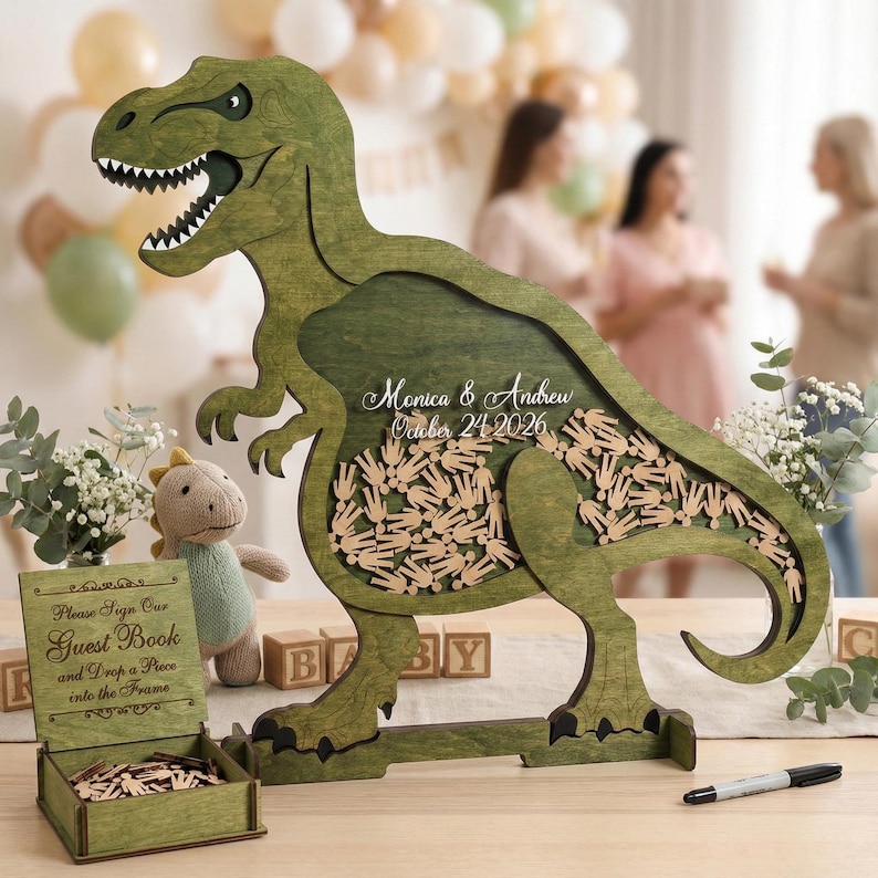 May include: A green wooden Tyrannosaurus Rex guest book alternative with a hollowed-out center filled with small wooden figures. The text "Monica & Andrew October 24 2026" is inscribed on the dinosaur. A small sign reads "Please Sign Our Guest Book and Drop a Piece into the Frame."
