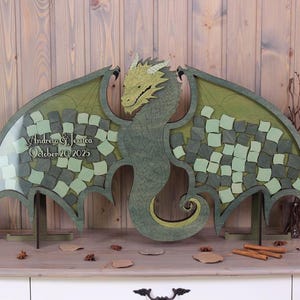 May include: Green wooden dragon wedding guest book with a green and white checkered design. The dragon has a large head and wings. The guest book is on a wooden stand. The text on the guest book reads "Andrew & Jessica October 20, 2025".