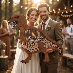 May include: A wooden dinosaur-shaped guest book with the names "Monica & Andrew" and the date "October 24 2026" engraved on it. The dinosaur has a large opening for guests to sign, and is held by a smiling couple at an outdoor wedding.