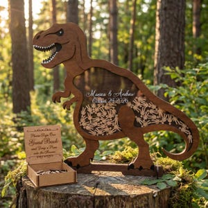 May include: A wooden Tyrannosaurus Rex guest book alternative with a slot for guests to drop wooden figures. The dinosaur has a clear center with the names "Monica & Andrew" and the date "October 24 2026". A small wooden box is included.
