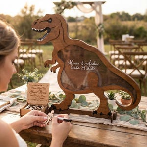 May include: A wooden dinosaur-shaped guest book frame with the names "Monica & Andrew" and the date "October 24 2026" inscribed. The frame has a clear window for a photo and a slot for guests to drop wooden pieces. A person is signing a wooden figure.