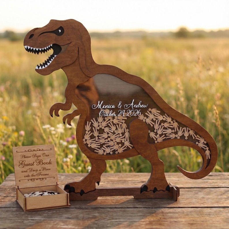 May include: A wooden Tyrannosaurus Rex-shaped guest book frame. The frame has a clear window filled with small wooden hand-shaped pieces. The text "Monica & Andrew October 24 2026" is written on the window. A small wooden box with the text "Please Sign Our Guest Book and Drop a Piece into the Frame" is in front.