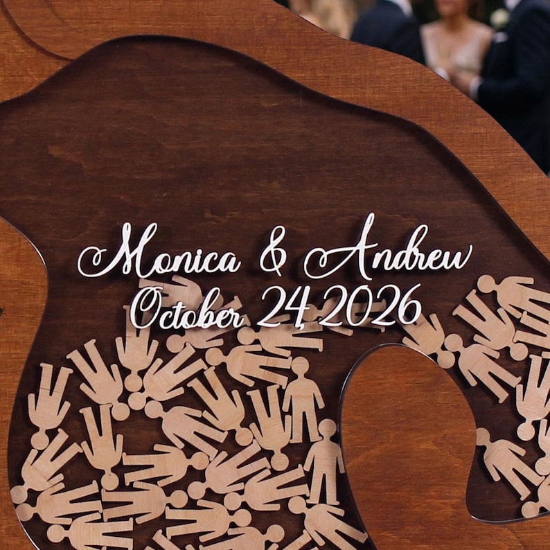 May include: Wooden wedding guest book alternative with the names "Monica & Andrew" and the date "October 24 2026" in white script. The design includes small wooden figures and a dark brown wood background.
