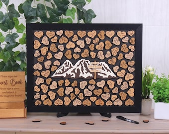 May include: A black rectangular frame containing a mountain cutout and numerous small wooden hearts attached to a dark background. The mountain features a wooden sign with the text "Izzy & Brendan" and "WILLIAMS". Each heart contains handwritten messages.