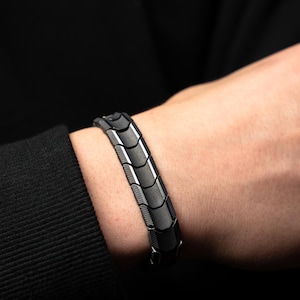 Op de afbeelding: Een close-up van een zwart en zilveren armband. De armband is gemaakt van in elkaar grijpende segmenten met een geometrisch ontwerp. De armband wordt om een pols gedragen, met een zwart shirt met lange mouwen op de achtergrond.