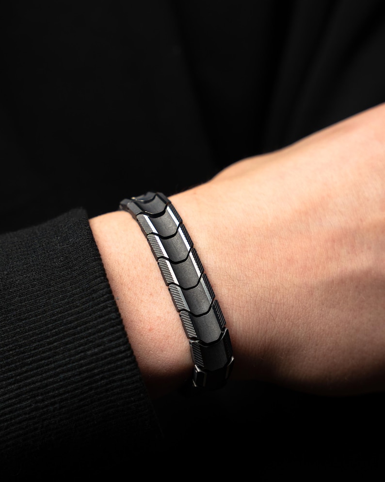 Op de afbeelding: Een close-up van een zwart en zilveren armband. De armband is gemaakt van in elkaar grijpende segmenten met een geometrisch ontwerp. De armband wordt om een pols gedragen, met een zwart shirt met lange mouwen op de achtergrond.