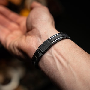 Op de afbeelding: Een zwarte armband met een geometrisch ontwerp wordt om een pols gedragen. De armband is gemaakt van rechthoekige schakels met een matte afwerking. Een zilveren accent is zichtbaar op sommige schakels. De armband heeft een sluiting met een driehoekig logo.