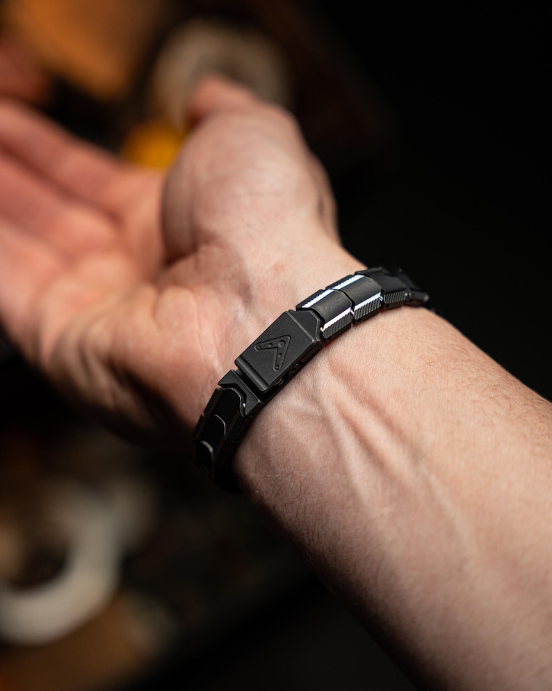 Op de afbeelding: Een zwarte armband met een geometrisch ontwerp wordt om een pols gedragen. De armband is gemaakt van rechthoekige schakels met een matte afwerking. Een zilveren accent is zichtbaar op sommige schakels. De armband heeft een sluiting met een driehoekig logo.