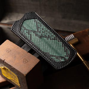 May include: A black leather wallet with a green snakeskin pattern. The wallet is stitched with black thread and has a smooth, polished finish.