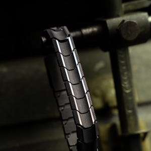 Könnte beinhalten: Ein schwarzes Metallarmband mit segmentiertem Design. Das Armband hat eine Reihe von gebogenen, rechteckigen Gliedern mit strukturierter Oberfläche und einem reflektierenden, silberfarbenen Rand. Das Armband wird vor einem dunklen, industriellen Hintergrund gezeigt.
