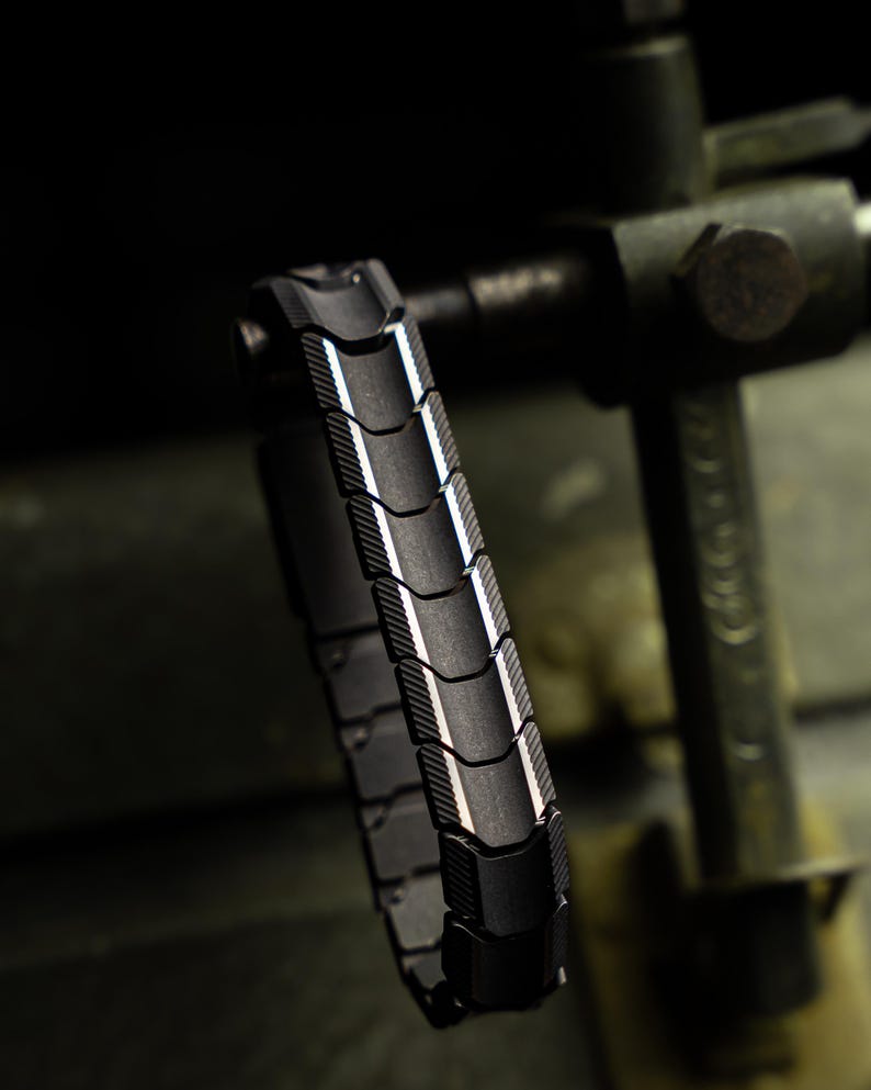 Op de afbeelding: Een zwartmetalen armband met een gesegmenteerd ontwerp. De armband heeft een reeks gebogen, rechthoekige schakels met een textuuroppervlak en een reflecterende, zilverkleurige rand. De armband wordt getoond tegen een donkere, industri&euml;le achtergrond.