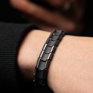 Op de afbeelding: Een zwarte armband met een geometrisch ontwerp wordt om een pols gedragen. De armband is gemaakt van verbonden, rechthoekige schakels met een matte afwerking en een subtiele glans. De achtergrond is donker, waardoor de details van de armband worden benadrukt.