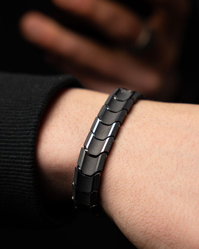 Op de afbeelding: Een zwarte armband met een geometrisch ontwerp wordt om een pols gedragen. De armband is gemaakt van verbonden, rechthoekige schakels met een matte afwerking en een subtiele glans. De achtergrond is donker, waardoor de details van de armband worden benadrukt.