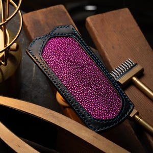 Puede incluir: Funda de cuero artesanal con un centro de piel de raya rosa vibrante, enmarcada por cuero negro y costuras azules. La funda descansa sobre tablas de madera, junto a una herramienta de latón y una lámpara de estilo vintage.