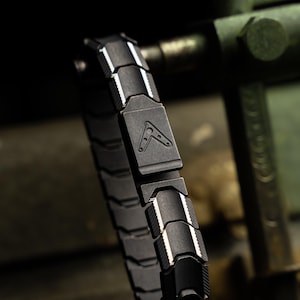 Op de afbeelding: Een zwart armband met een geometrisch ontwerp. De armband is gemaakt van in elkaar grijpende segmenten met witte accenten en een centrale sluiting met een logo. De armband wordt getoond tegen een vage, industri&euml;le achtergrond.
