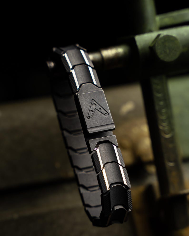 Op de afbeelding: Een zwart armband met een geometrisch ontwerp. De armband is gemaakt van in elkaar grijpende segmenten met witte accenten en een centrale sluiting met een logo. De armband wordt getoond tegen een vage, industri&euml;le achtergrond.