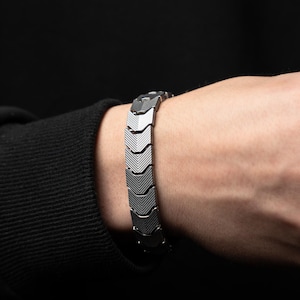 May include: A silver bracelet with a geometric chevron pattern. The bracelet is composed of linked segments, each with a textured design. The bracelet is worn on a wrist against a dark background.