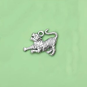 May include: Silver charm in the shape of a tiger. The tiger is depicted in a crouching position with its head turned to the side. The charm has a loop at the top for attaching to a bracelet or necklace.
