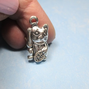 May include: Silver charm of a Japanese Maneki-Neko, or lucky cat, with its paw raised. The charm has a raised inscription on the paw that reads "Good Luck".