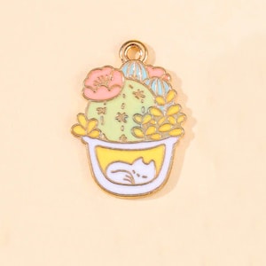 May include: A gold-tone metal charm featuring a cartoon cactus in a white pot with a yellow interior. The cactus is green with pink, yellow, and blue flowers and leaves. A white cat is sleeping inside the pot.