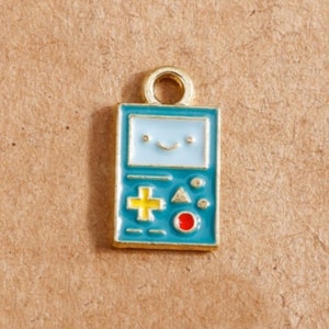 May include: A rectangular charm with a gold loop at the top. The charm features a teal background with a smiling face, a yellow cross, and other colorful details. The charm is likely for jewelry or crafting.