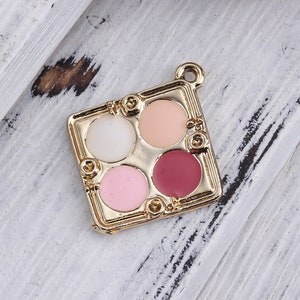 May include: Gold-toned metal charm shaped like a makeup palette with four round compartments. The compartments are filled with pink, white, peach, and burgundy colored enamel.