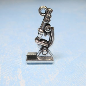 May include: Silver charm in the shape of a microscope. The microscope has a round base and a long, thin tube with a lens at the top.
