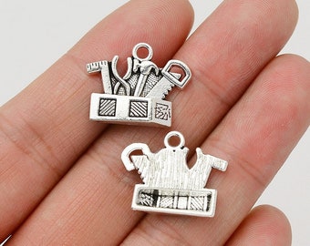 May include: Two silver charm pendants, one with a toolbox and tools, and the other with a teapot and teacups. Both charms have a textured surface and are in the shape of a box.