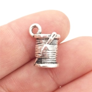 May include: Silver charm in the shape of a spool of thread with a needle sticking out of it. The charm has a loop at the top for attaching to a necklace or bracelet.