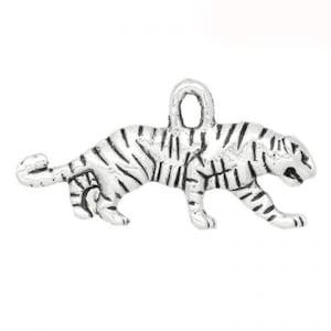 May include: Silver charm in the shape of a tiger. The tiger is depicted in a walking pose with its head turned to the side. The charm has a loop at the top for attaching to a necklace or bracelet.