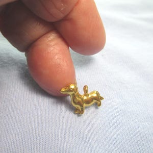 May include: A small gold-colored dachshund charm pendant. The charm is detailed and has a loop at the top for attaching to a necklace or bracelet.