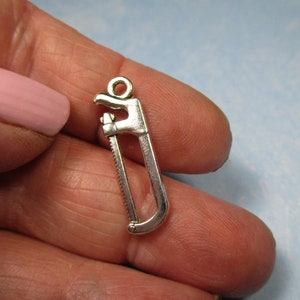 May include: Silver-toned saw charm pendant. The charm is shaped like a hand saw with a loop at the top for attaching to a chain or bracelet.