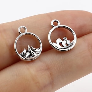 May include: Two silver charm pendants with a circular design. One pendant features a mountain range inside the circle, and the other pendant features a wave inside the circle.