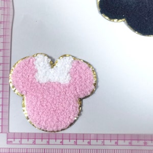 May include: Pink and white fuzzy fabric applique in the shape of a mouse head with a gold outline. The applique is about 5 cm wide.