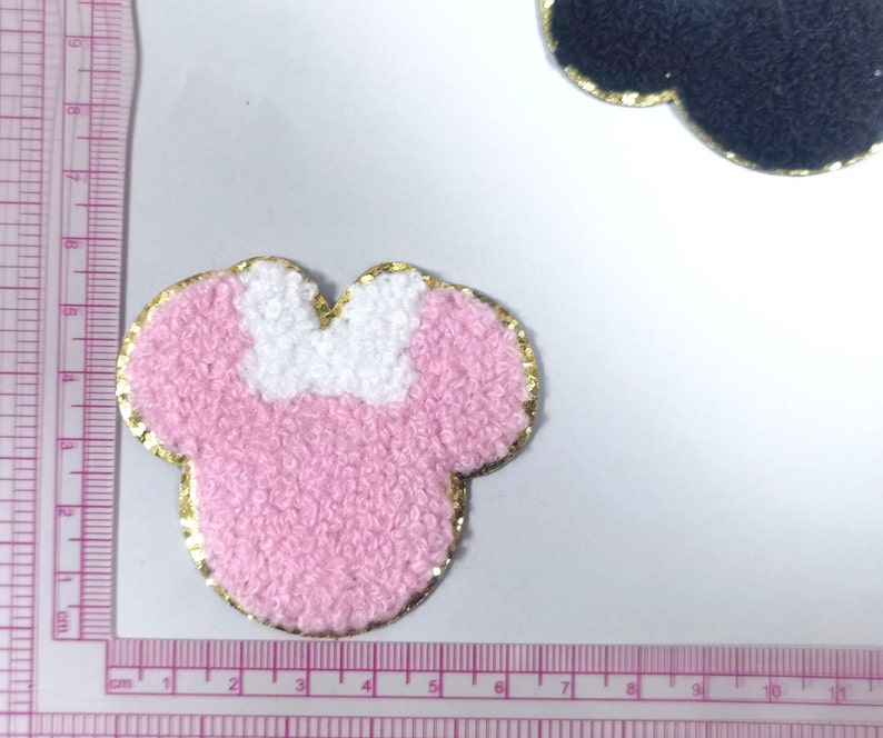 May include: Pink and white fuzzy fabric applique in the shape of a mouse head with a gold outline. The applique is about 5 cm wide.