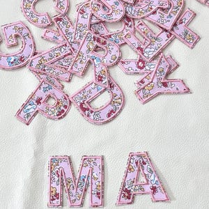 K&ouml;nnte beinhalten: Rosa Stoffbuchstaben mit Blumenmuster. Die Buchstaben ergeben "MA".
