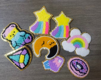 May include: A collection of colorful, embroidered patches with gold glitter borders. Designs include a star with a rainbow, a toucan, a rainbow, a donut, and other whimsical shapes. These patches are likely for clothing or accessories.
