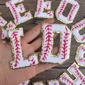 Pode incluir: Letras de feltro branco com acabamento dourado brilhante e costuras vermelhas, que se assemelham a uma bola de beisebol. As letras soletram "LD".