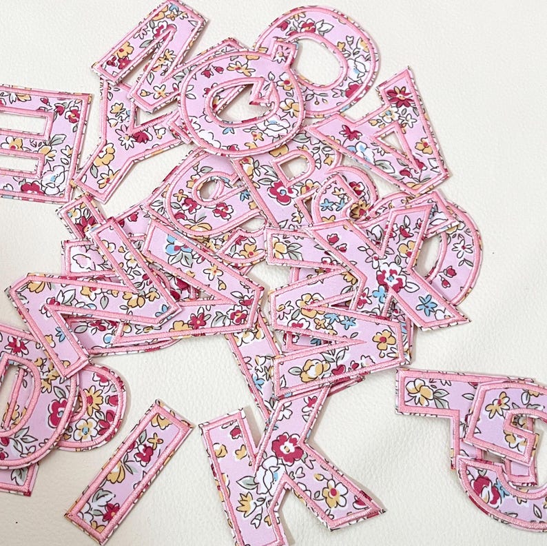 K&ouml;nnte beinhalten: Eine Sammlung von pinkfarbenen Stoffbuchstaben mit einem Blumenmuster. Die Buchstaben sind mit einem dunkleren Pink umrandet und haben einen wei&szlig;en Hintergrund.